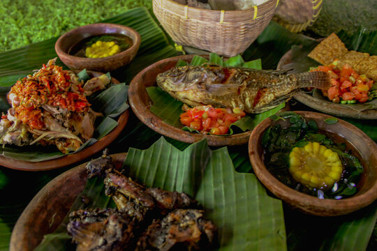 Traditional sasak dish consisting of ayam merangkat, fried tilapia, fried chicken, fried tempe with sambal, clear vegetable soup, and rice served on a clay plate and banana leaves.