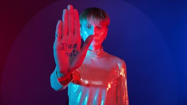 Young adult woman showing palms with affirmation I am not victim in colorful lighting indoors. Create own reality. Female person short hair wearing sparkling reflective clothing. Social issue concept
