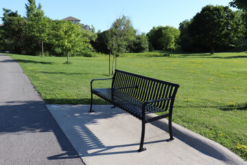 Fototapeta na wymiar Stand-alone black metal bench in a park on a walkway surrounded with green grass lawn and trees on a sunny Summer day