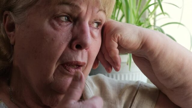 Old woman crying frantically at home. Elderly wrinkled grandmother wipes her tears with hand. Pain, disappointment, depression, loneliness, old age 