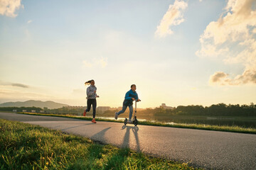 Obraz premium Happy mother and son go in sports outdoors. Boy rides scooter, mom runs on sunny day. Silhouette family at sunset. Health care, authenticity, sense of balance and calmness. 