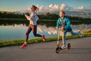 Happy mother and son go in sports outdoors. Boy rides scooter, mom runs on sunny day. Silhouette family at sunset. Health care, authenticity, sense of balance and calmness.
