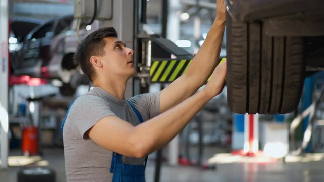 Side view concentrated Caucasian young handsome man examining car tyre indoors. Focused professional expert maintenance engineer repairing automobile at service station
