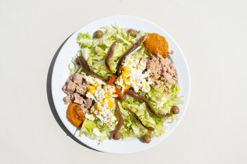 esquerola salad with tuna, anchovies, olives, grated hard-boiled egg and romesco sauce. white plate on a white background with warm sunlight for a fresh and vitamin salad.