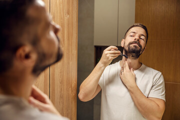 Young bearded man is shaving his beard with razor blade in front of the mirror in a bathroom.