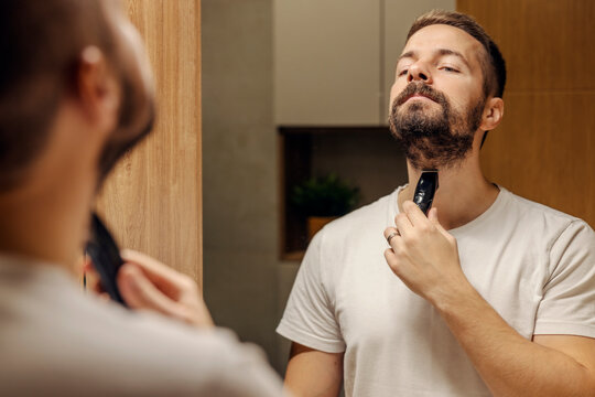 Reflection of a man trimming his beard in a bathroom in front of the mirror.
