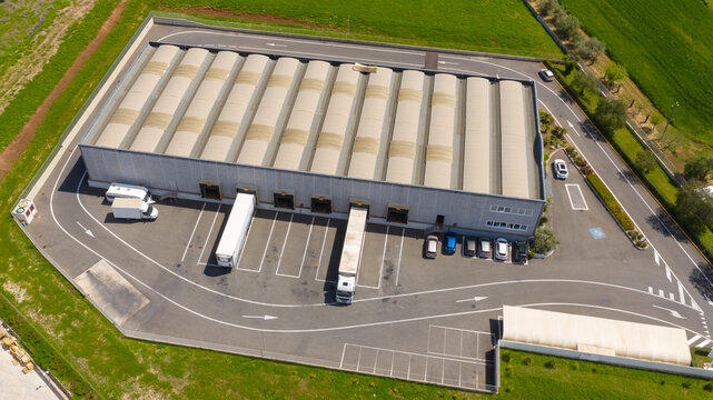 Aerial view of a large shed that is used as a warehouse for loading and unloading parcels. Many trucks are parked around the facility for their work. All within an industrial area.
