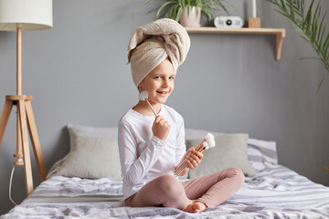 Smiling happy positive cute little girl holding makeup brushes, wearing white towel sitting in bed in bedroom morning routine procedures playing with mother cosmetics tools.