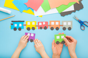 Mother and child hands creating beautiful colorful paper train with wagons on light blue table background. Pastel color. Point of view shot. Closeup. Playing and spending time together. Top down view.