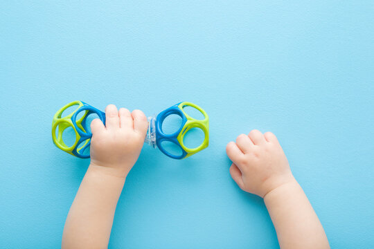 Baby boy hands playing with colorful rattle dumbbell on light blue table background. Pastel color. Closeup. Point of view shot. Toy of development for infant. Top down view.