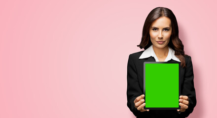 Obraz premium Portrait of serious brunette businesswoman showing tablet pc, touchpad, with green chroma key screen, on rose pink background. Confident attractive business woman at office.