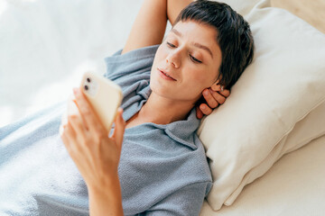 A young woman lying on the bed uses a mobile phone for social networks and online messaging.