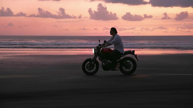 Happy motorcyclist ride motor bike. Brutal biker enjoy epic pink sunset view. Male person move motorbike. Rider man drive motorcycle. Sun set sea shore. Beach moto trip slow motion. Guy ocean travel.