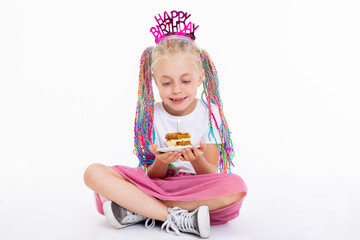 Delighted preschooler daughter kid with funny braids wearing white t-shirt and pink skirt and funny colorful braids holding plate with delicious tasty piece of cake with candle posing in white studio.