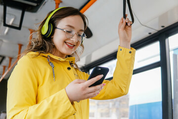 Smiling young woman in yellow jacket wearing headphones standing on a bus listening to music and...