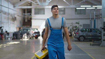 Satisfied Caucasian young man with tool kit box strolling at service station smiling. Dolly shot front view portrait of happy confident handsome mechanic walking in slow motion indoors