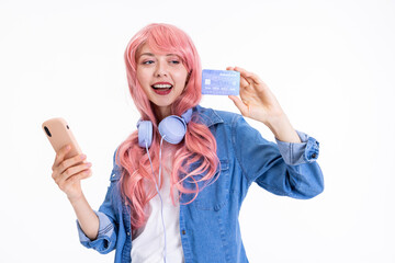 Smiled young woman holding credit card and smartphone in hands showing at camera wearing pink wig and modern headphones on neck posing for camera advertisiment of bank.