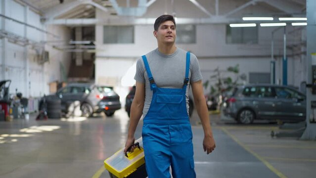 Satisfied Caucasian young man with tool kit box strolling at service station smiling. Dolly shot front view portrait of happy confident handsome mechanic walking in slow motion indoors