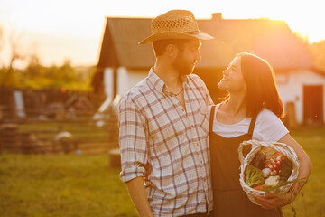 Portrait of young happy farmer couple holding basket with fresh organic vegetables on their rancho at the sunset. Vegetarian healthy food. Family business. Ecological farm and agriculture business.