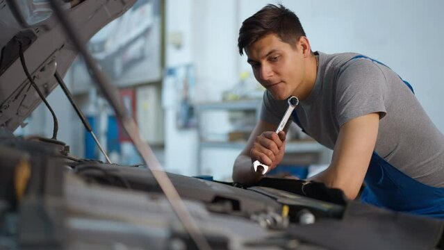 Side view portrait of concentrated thoughtful mechanic leaning on car hood touching face with wrench. Unsure Caucasian young man in uniform thinking on broken automobile repair at car service station
