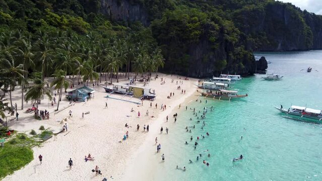 Aerial: Filipino Outrigger Tour Boats anchored on Tropical Seven Commandos Beach, Tourists swimming in turquoise clear water on white sand, Palm trees and lush green limestone mountains.El Nido