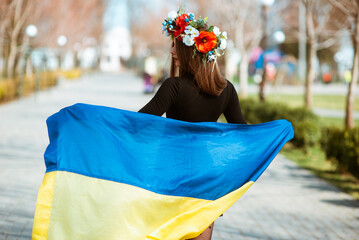 young woman holds in hands flag of Ukraine with wreath of flowers on head standing back. Patriotic...