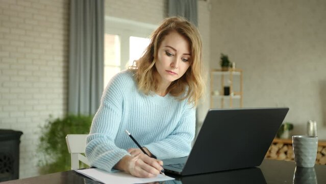 Young blonde is making notes on sheet of paper. The woman prints and searches for information in notebook. Beautifully furnished room in the background, High quality 4k footage