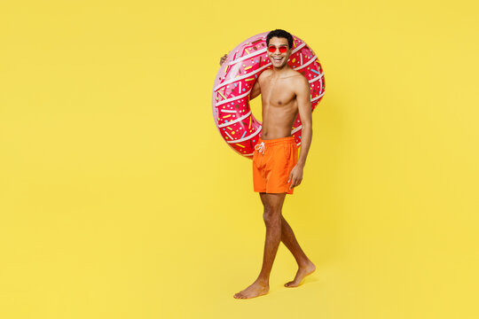 Full body happy fun young man wear orange shorts swimsuit relax near hotel pool hold pink donut rubber ring walking going isolated on plain yellow background Summer vacation sea rest sun tan concept.