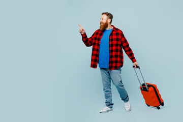 Traveler man wear casual clothes hold suitcase point aside go isolated on plain pastel blue background studio. Tourist travel abroad in free spare time rest getaway. Air flight trip journey concept.