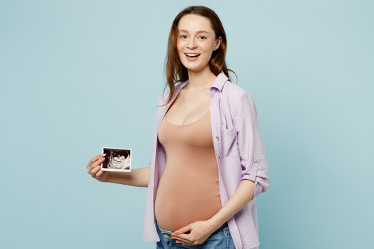 Sideways young pregnant future mom woman with belly tummy wearing casual clothes hold show ultrasound image pregnant baby photo isolated on plain pastel blue background. Maternity pregnancy concept.