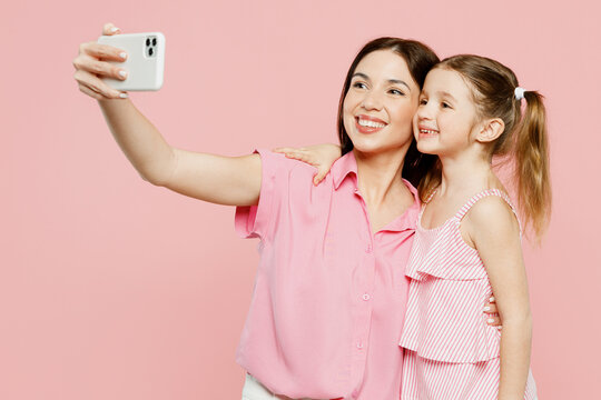 Side view happy woman wearing casual clothes with child kid girl 6-7 years old. Mother daughter do selfie shot on mobile cell phone isolated on plain pastel pink background. Family parent day concept.