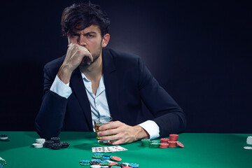 Gambling Concepts. Thoughful Handsome Caucasian Brunet Cards Player At Pocker Table With Chips and Cards While Playing and Drinking Alcohol.
