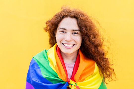 Portrait of red haired non binary man with rainbow flag smiling at camera over yellow background. LGBT community, diversity, transgender and non-binary people concept