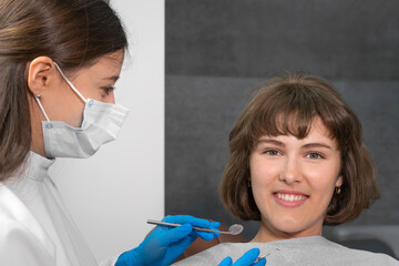 Young smiling woman is sitting in a dental chair and looking into the camera, the dentist is...