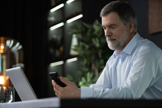 Smiling mature businessman holding smartphone sitting in office. Middle aged manager ceo using cell phone mobile apps and laptop. Digital technology applications and solutions for business development
