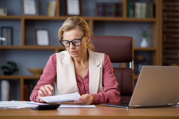 Portrait of adult businesswoman wearing eyeglasses working at office. Female manager doing paperwork, analyzing financial report, holding documents with graphs and charts at workplace