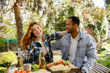 Two friends are having fun on a picnic, telling jokes, drinking drinks. A redhaired girl and an African American oy are having fun outside the city and in the company of friends.