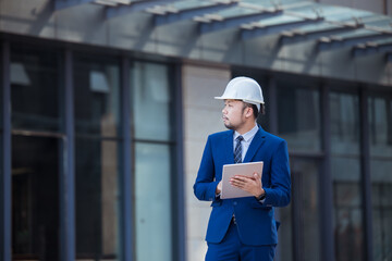 Handsome engineer wearing suit and hard hat or white helmet on building background. Engineer using tablet for checking construction about modern building. copy space for text.