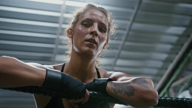 Woman fighter leaning on ring ropes in gym. Portrait of female tired boxer covered in sweat recovers breath while training and looking at camera