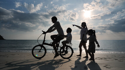Fototapeta premium Asian family On Summer Vacation Four people Father, mother, daughter and son riding bicycles and walking In the evening the sunset on the beach. Travel and Relaxation