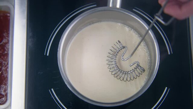 Chef cook keeps spiral whisk in hand and waits boiling cream sauce in stewpan on electric stove. Process of making delicious dishes. Concept of working on restaurant kitchen. Slow motion. Close up.