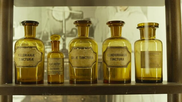 Antique glass jars with ancient medicines stand on wooden shelf with warm lighting. Shabby labels with printed names of homeopathic remedies in Latin, no people.