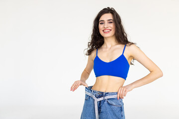 Young woman measuring her waist and showing how much weight she lost in jeans, on a white background.
