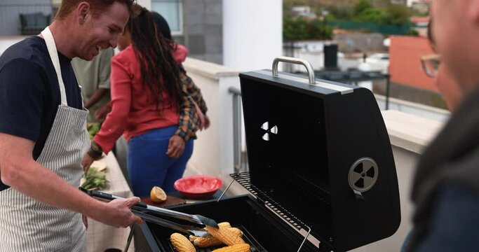 Multi generational people doing barbecue during weekend day at home's rooftop - Multiracial friends having fun cooking together corn cobs on the girll - Summer and food concept