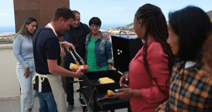 Multi generational people doing barbecue during weekend day at home's rooftop - Multiracial friends having fun together putting corn cobs on the girll - Summer and food concept