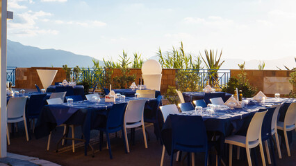 Restaurant, cafe on outdoor terrace with view of sky and mountains. Tables set with blue linen and white dishes.