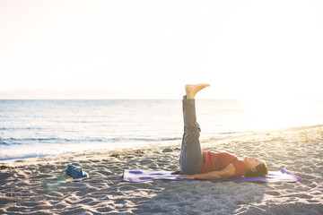 Senior woman exercising, stretching on yoga mat at the beach, doing leg raises.