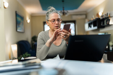 grandmother wearing glasses sitting at desk with laptop holding mobile, reading text message, chatting with grandchild