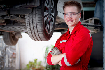 Portrait of smiling handsome mechanic in red uniform work with lifted vehicle, auto mechanic standing arms crossed next to wheel, technician repair and maintenance customer car automobile at garage.