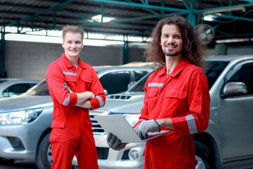 Handsome mechanic men in red uniform holding laptop computer standing at vehicle garage, auto mechanic technician teamwork working together, repairing customer car automobile at car service shop.